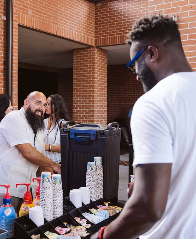 Two men at a coffee station with cups, sugar packets, and creamers, inside a brick building with other people in the background.