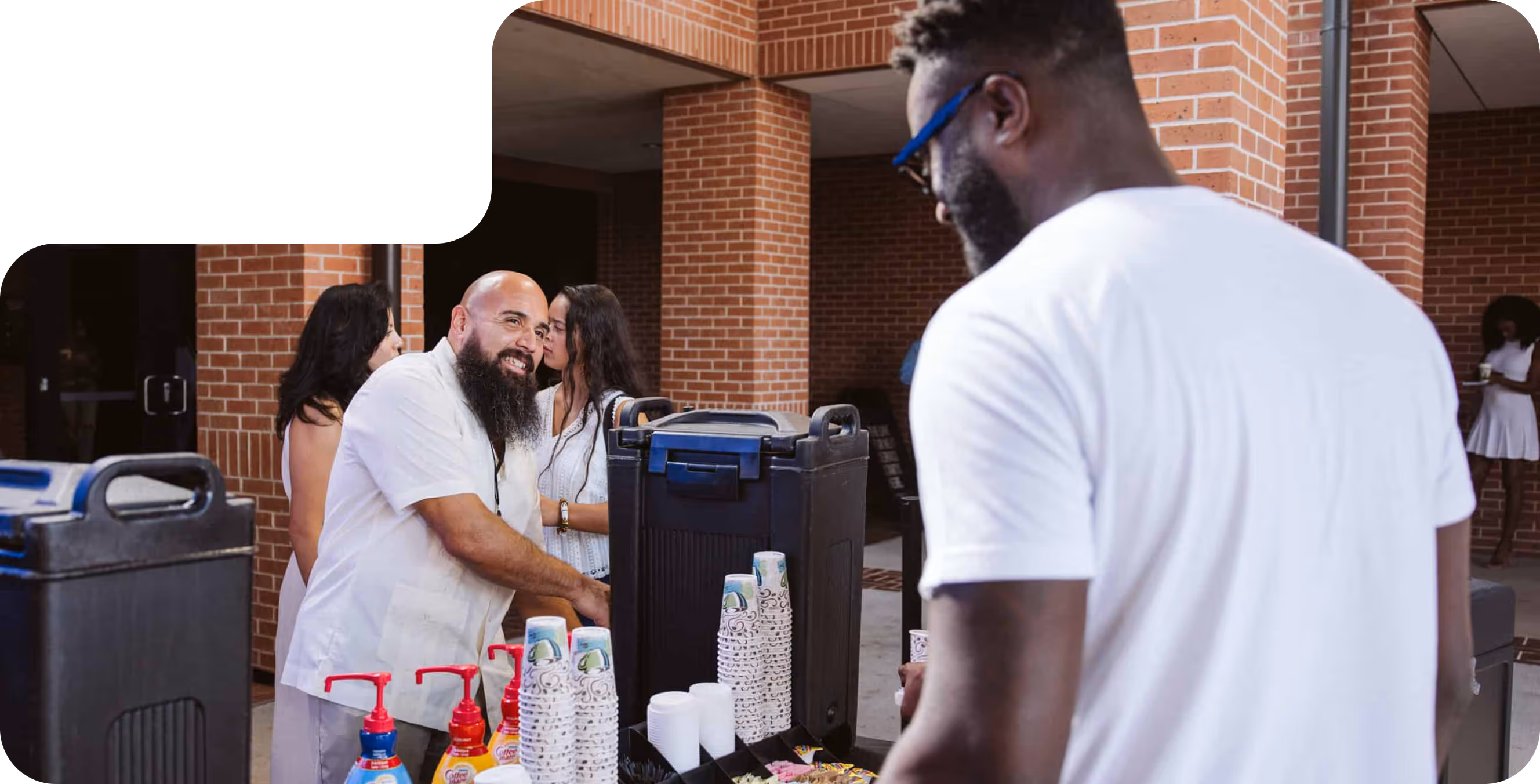 Two men at a coffee station with cups, sugar packets, and creamers, inside a brick building with other people in the background.