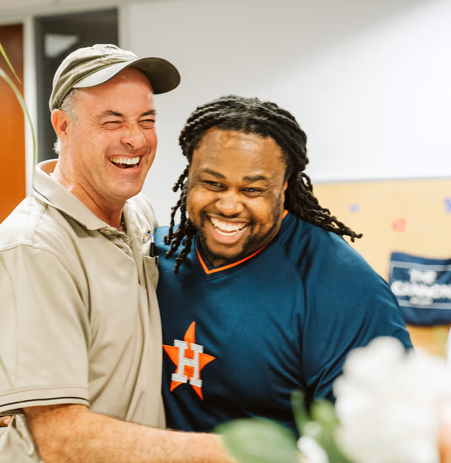 Two men smiling and hugging indoors, one wearing a beige cap and polo shirt, the other in a Houston Astros jersey.
