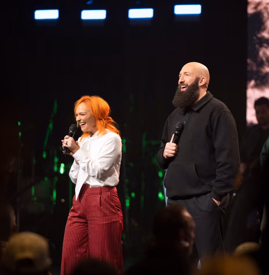 A smiling woman with orange hair in a white blouse and red pants and a bearded man in a black sweater hold microphones while speaking on stage.