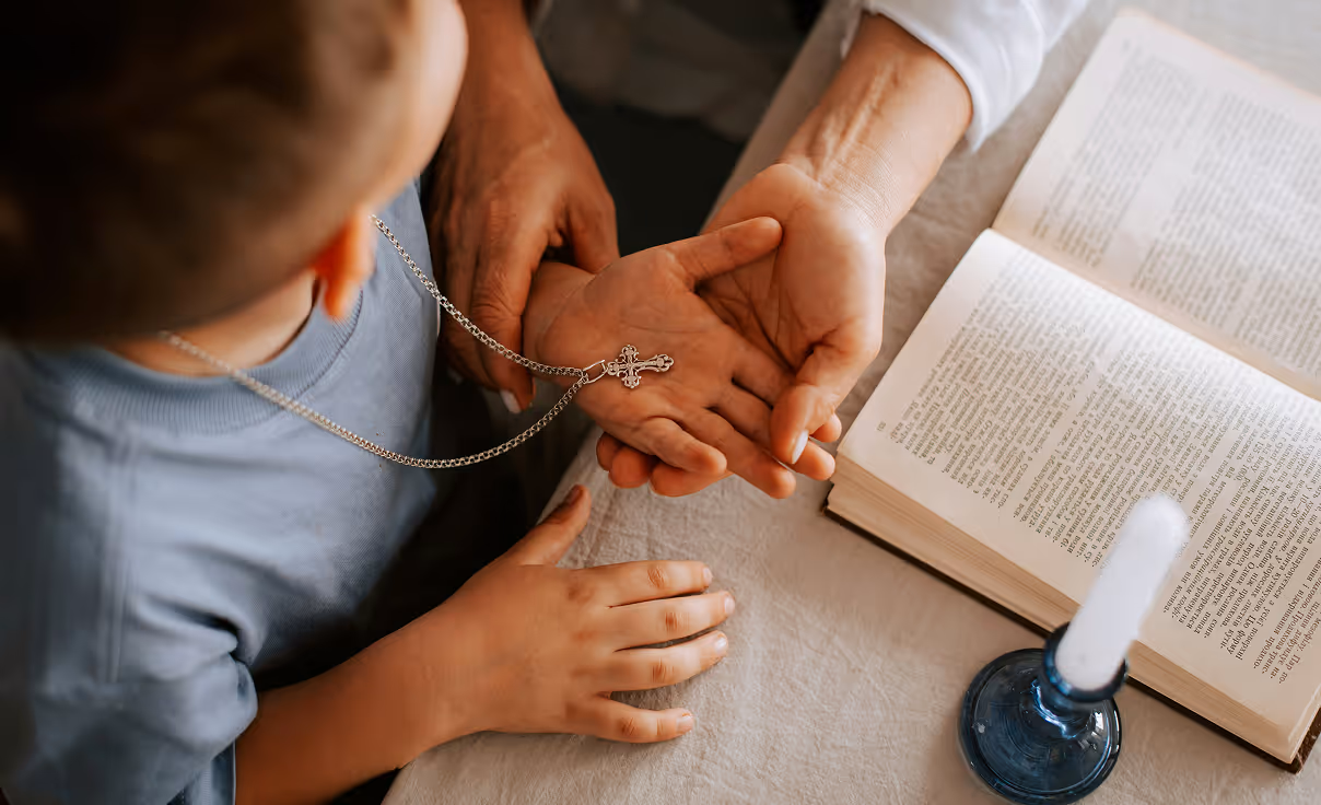 Child holding a silver cross necklace in adult's hands next to an open book and a candle on a table.