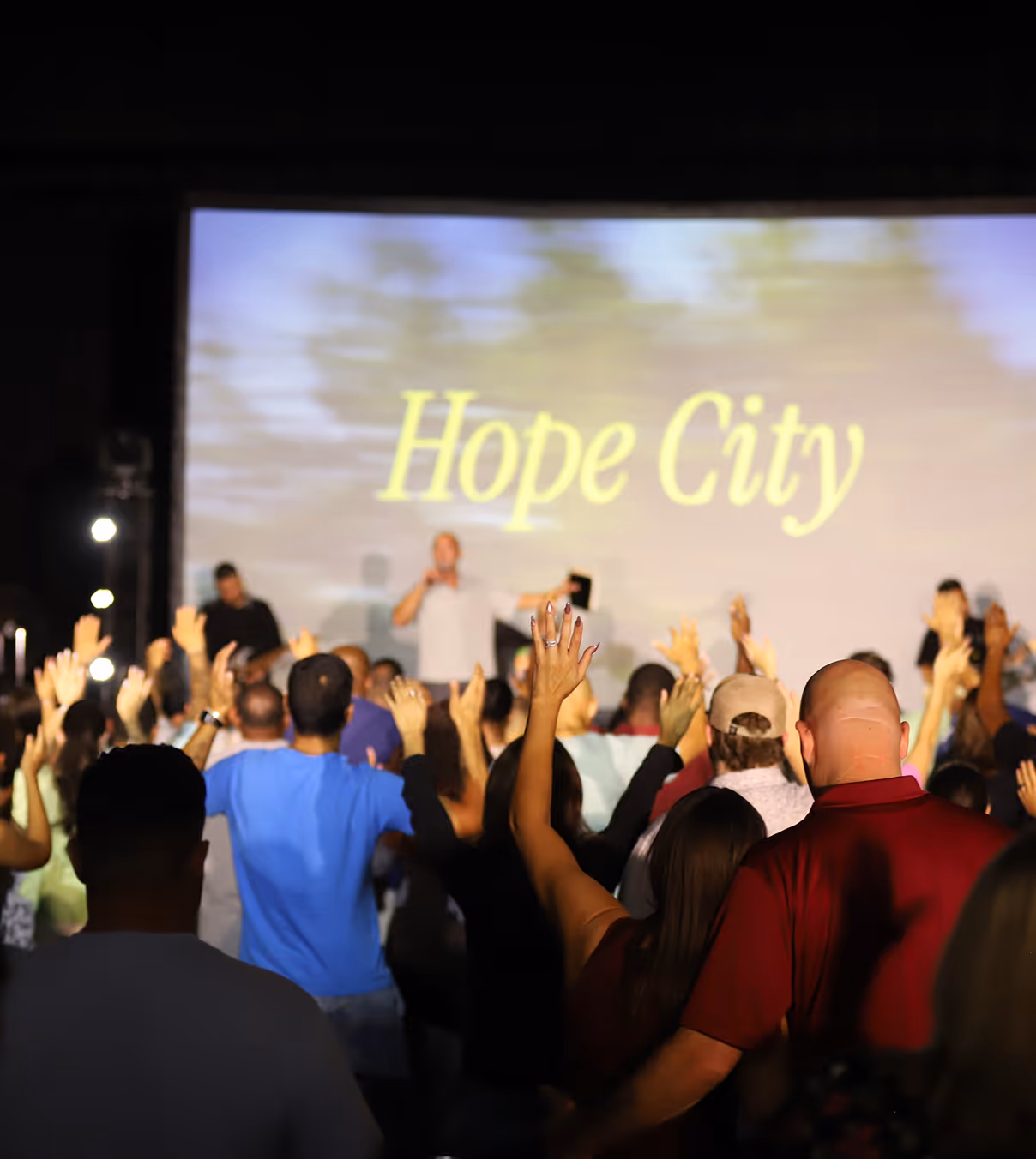 Crowd with raised hands facing a stage where a speaker stands in front of a screen displaying the words 'Hope City'.