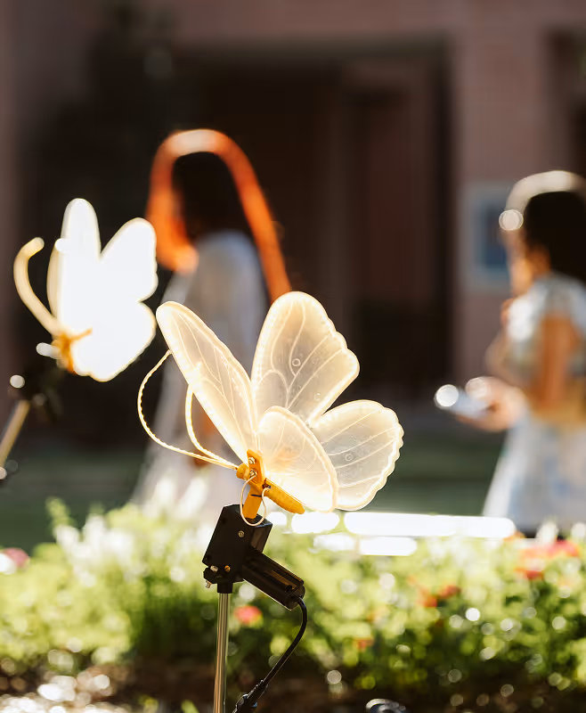 Glowing decorative butterfly lights on stakes with blurred people and flowers in the background.