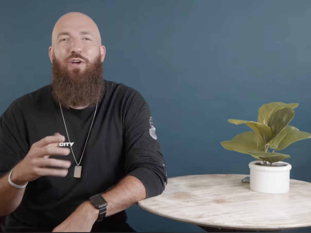 Bearded man in black shirt gesturing with one hand while sitting at a round wooden table with a potted plant against a blue background.