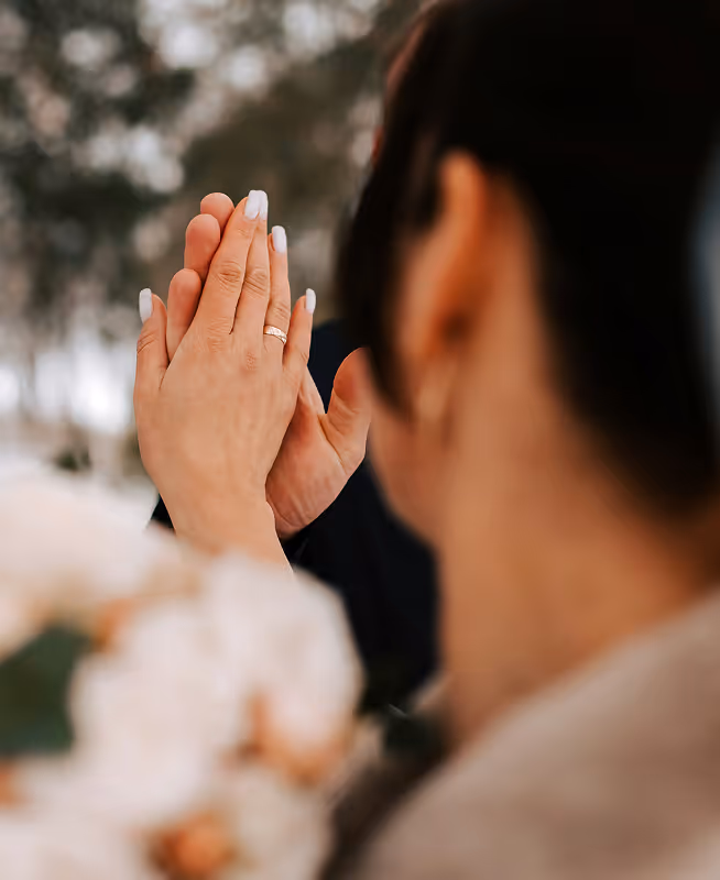 Close-up of two people touching hands with a visible wedding ring, blurred bouquet and background.
