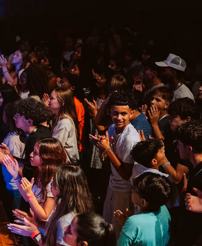 A diverse group of young people standing closely together and clapping in a dimly lit environment.