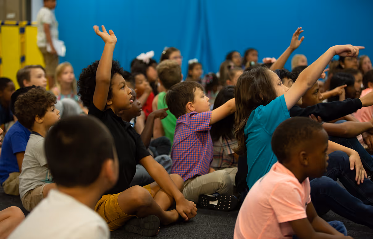 Group of diverse young children sitting on the floor in a classroom, several raising hands to participate.