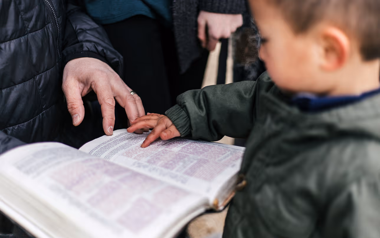 An adult and a child pointing at the text in an open book, possibly a Bible.