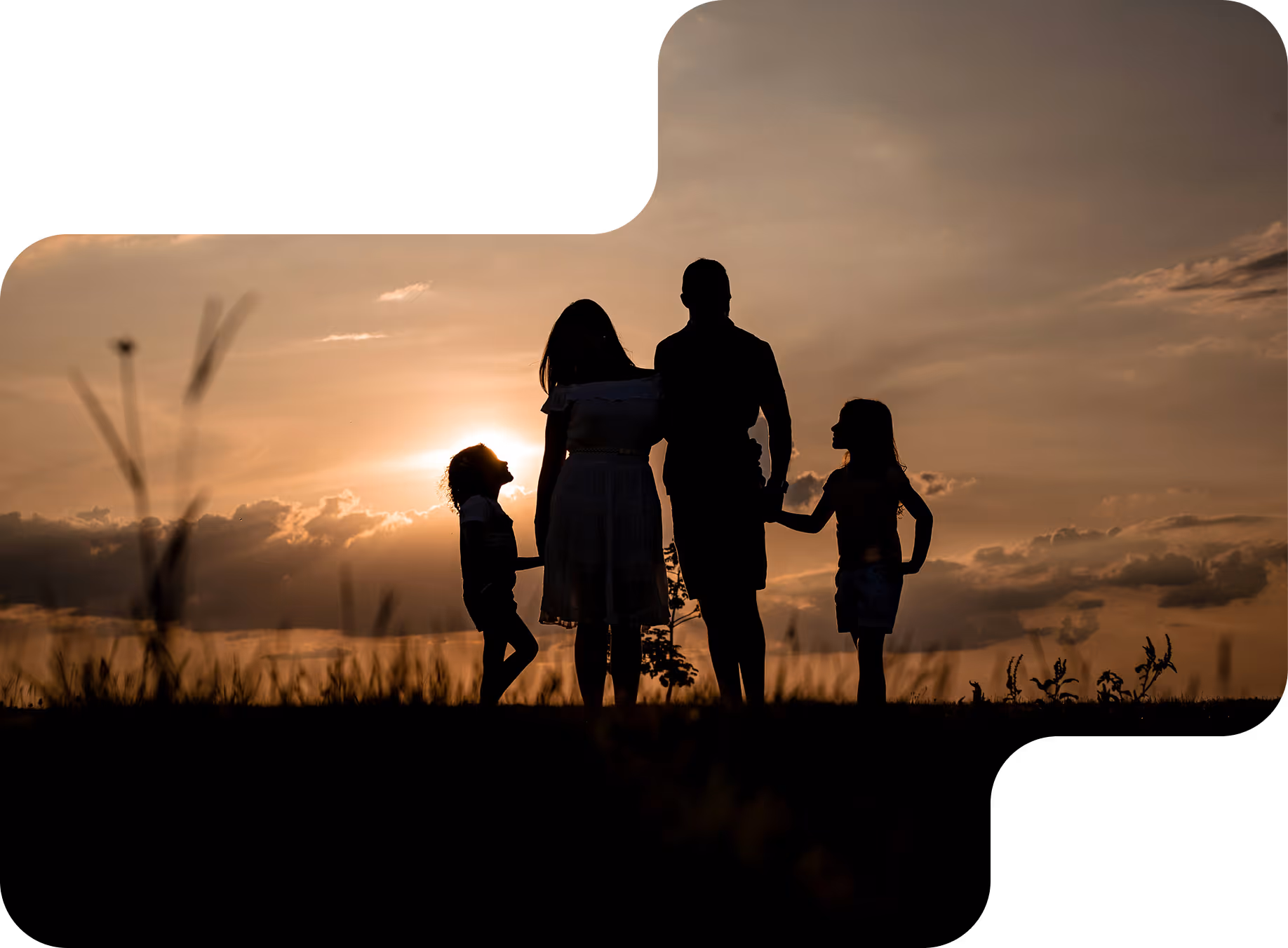 Silhouette of a family of four holding hands outdoors at sunset with a cloudy sky.