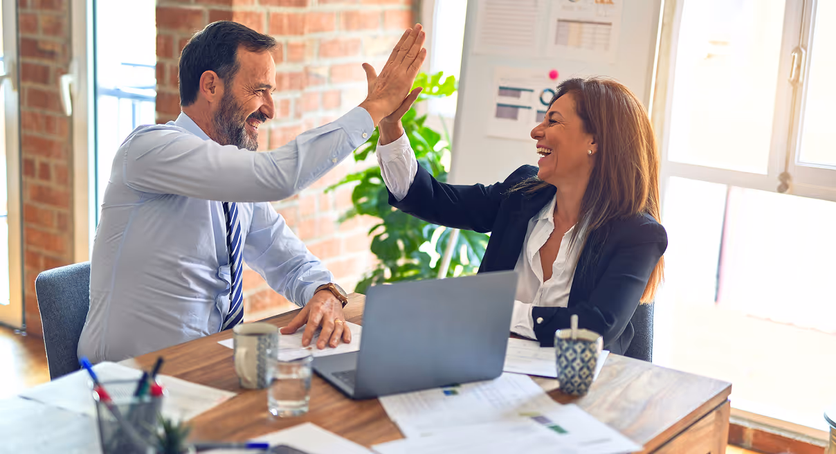 A man and a woman in business attire smiling and giving each other a high five across a desk with a laptop and documents.