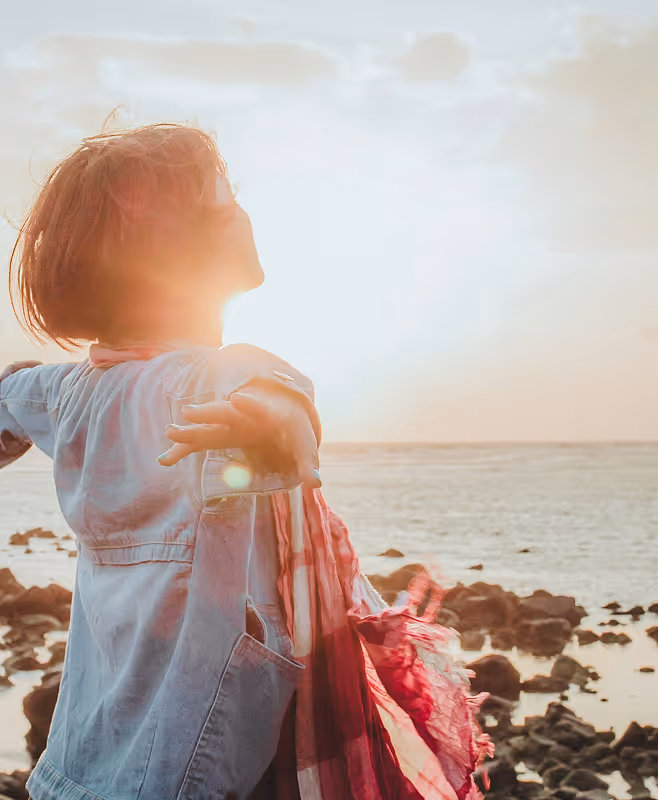 Person standing on rocky shore with arms outstretched facing the sun over the ocean at sunset.