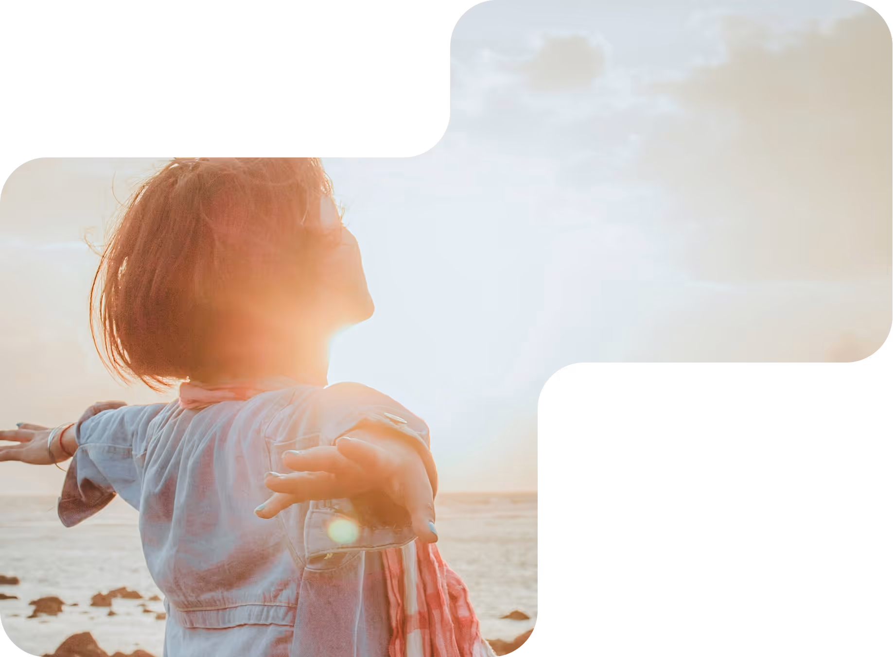 Person standing on rocky shore with arms outstretched facing the sun over the ocean at sunset.