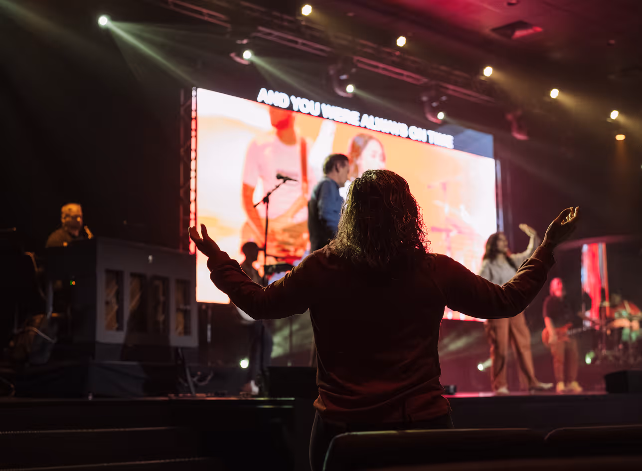 Person with raised arms worshiping during a live music performance on a stage with a large screen in the background.