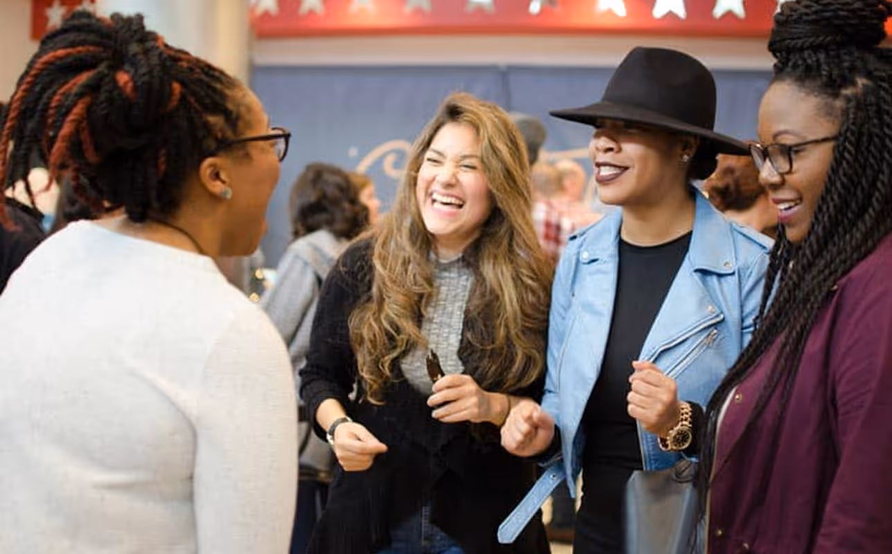 Four women happily chatting and laughing together indoors, one wearing a black hat and light blue jacket.