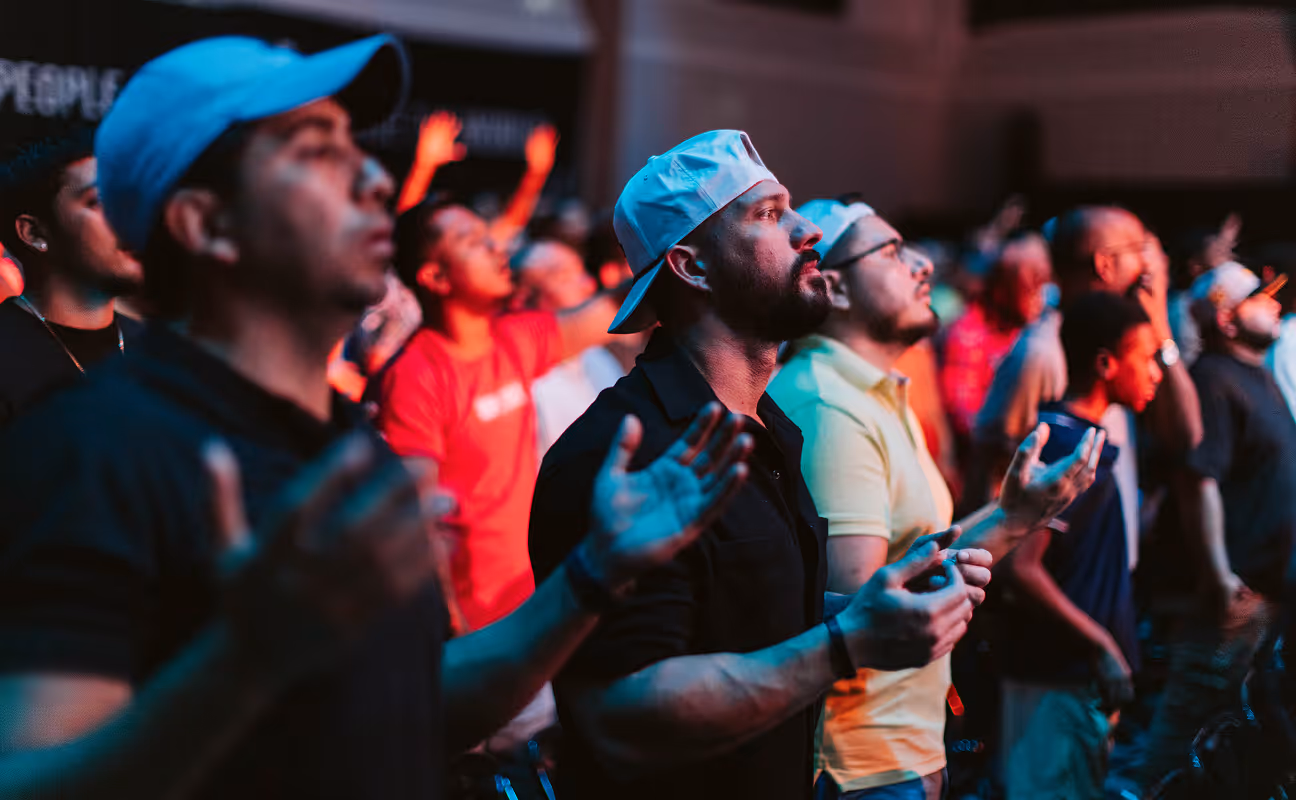 Group of men standing with eyes closed and hands raised, engaged in prayer or worship at an indoor event.