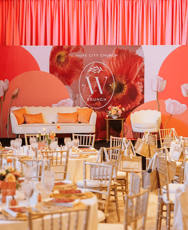 Event setup with gold chairs and round tables in front of a stage with white couch, orange pillows, floral arrangement, and a backdrop reading 'Hope City Church W Brunch'.
