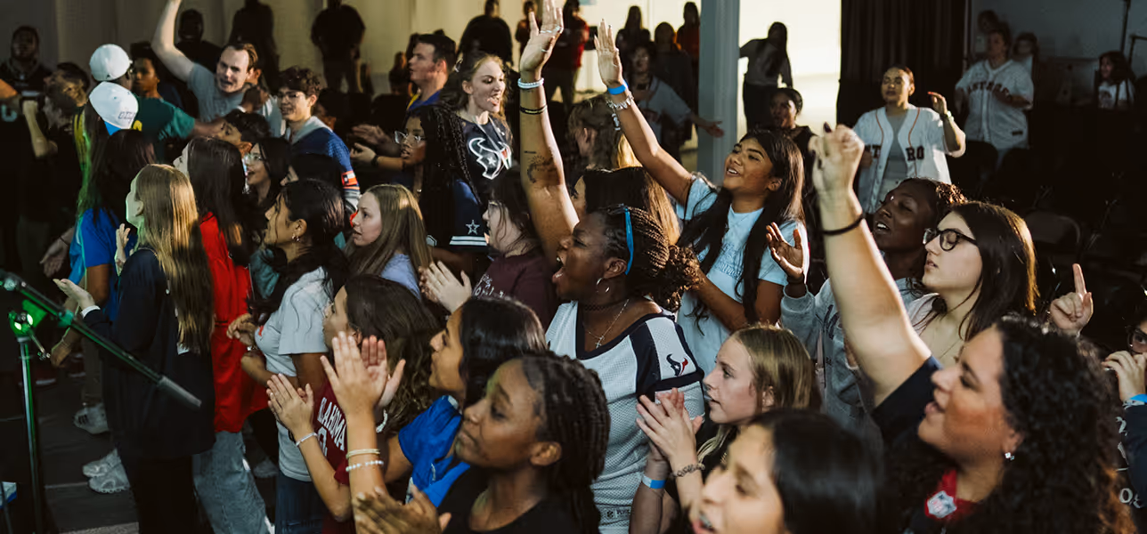 Diverse group of young people standing closely, raising hands and clapping in an indoor gathering.