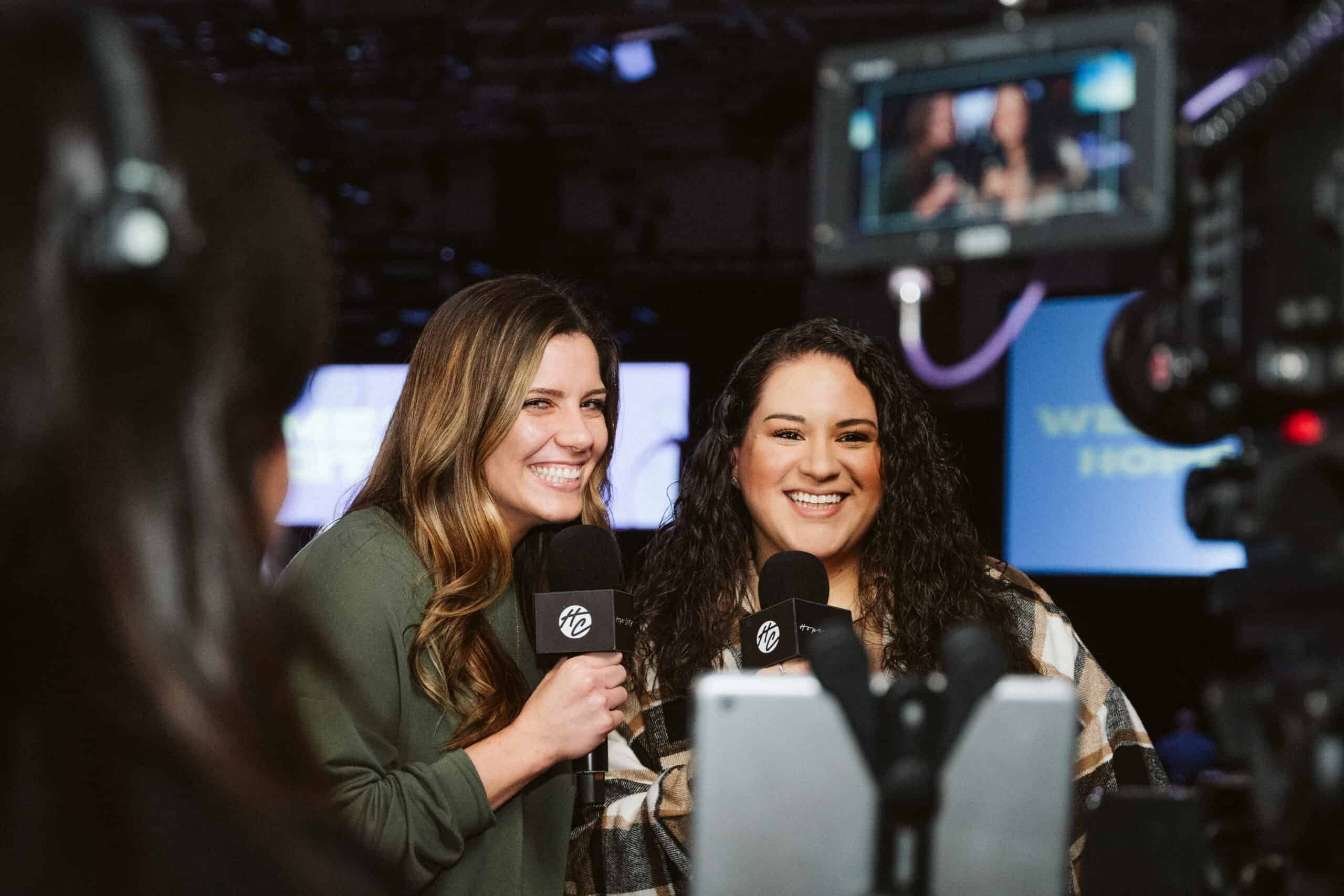 Two women smiling and holding microphones in front of cameras during a recording session.