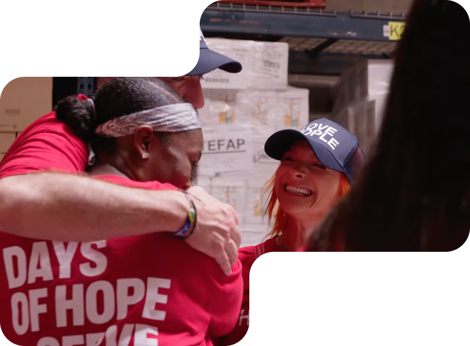 Three volunteers wearing red and blue shirts and hats hugging and smiling in a warehouse.
