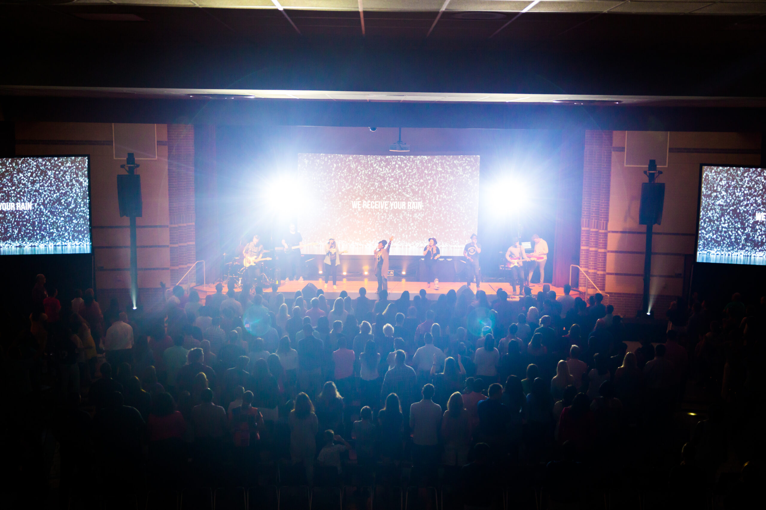 A band performing on stage in front of a large audience with bright lights and screens displaying text.