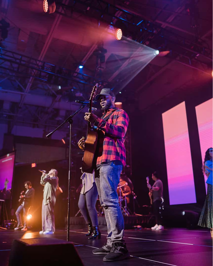 Musician in red plaid shirt playing guitar on stage with band under colorful stage lights.