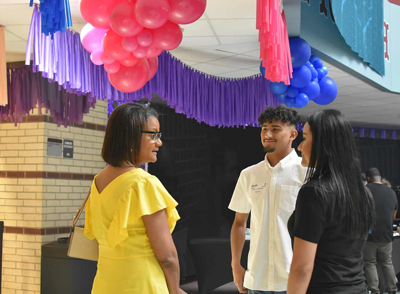 Three people talking indoors under colorful hanging balloon and paper decorations.