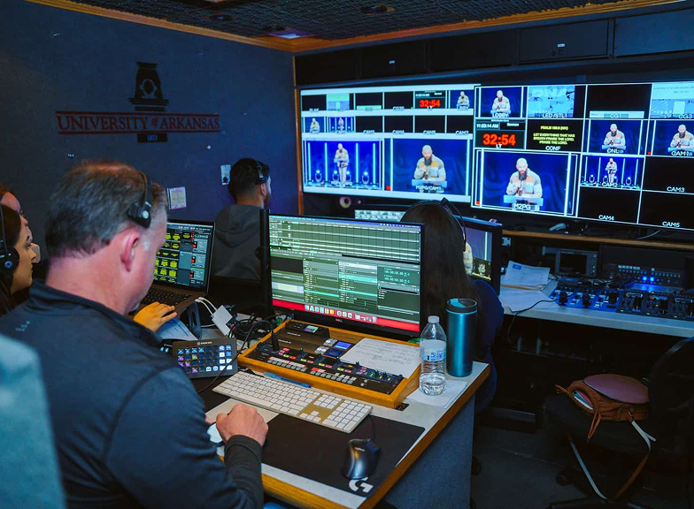 People with headsets working at control panels in a video production room with University of Arkansas logo.