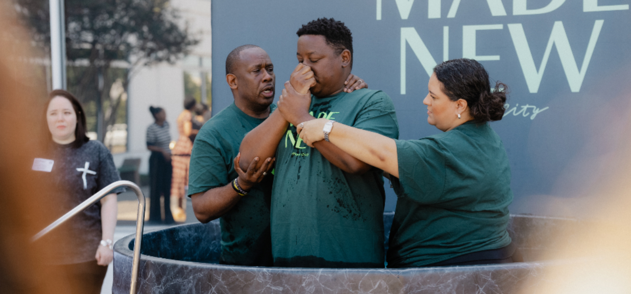 Smiling man with raised fist wearing wet blue shirt labeled I WILL FOLLOW being supported by another man in a DREAM TEAM shirt during baptism.