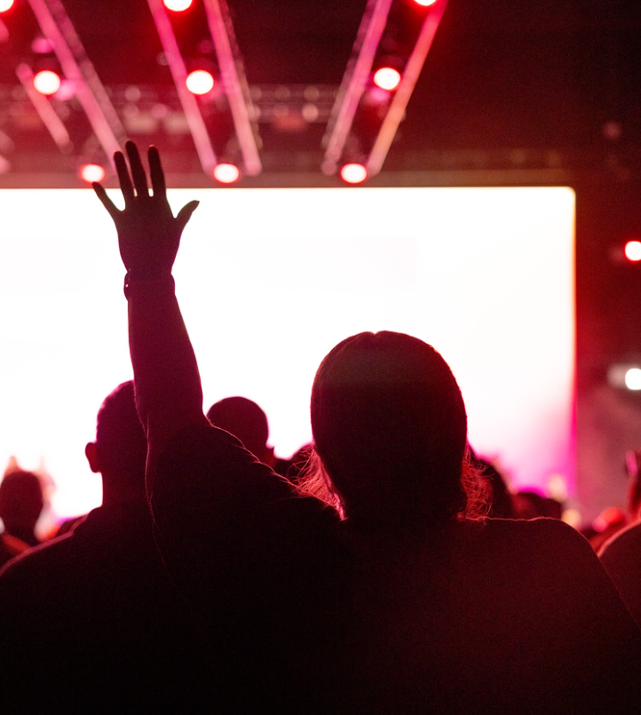 Live worship band performing on stage with audience watching under colorful lights and screen text.