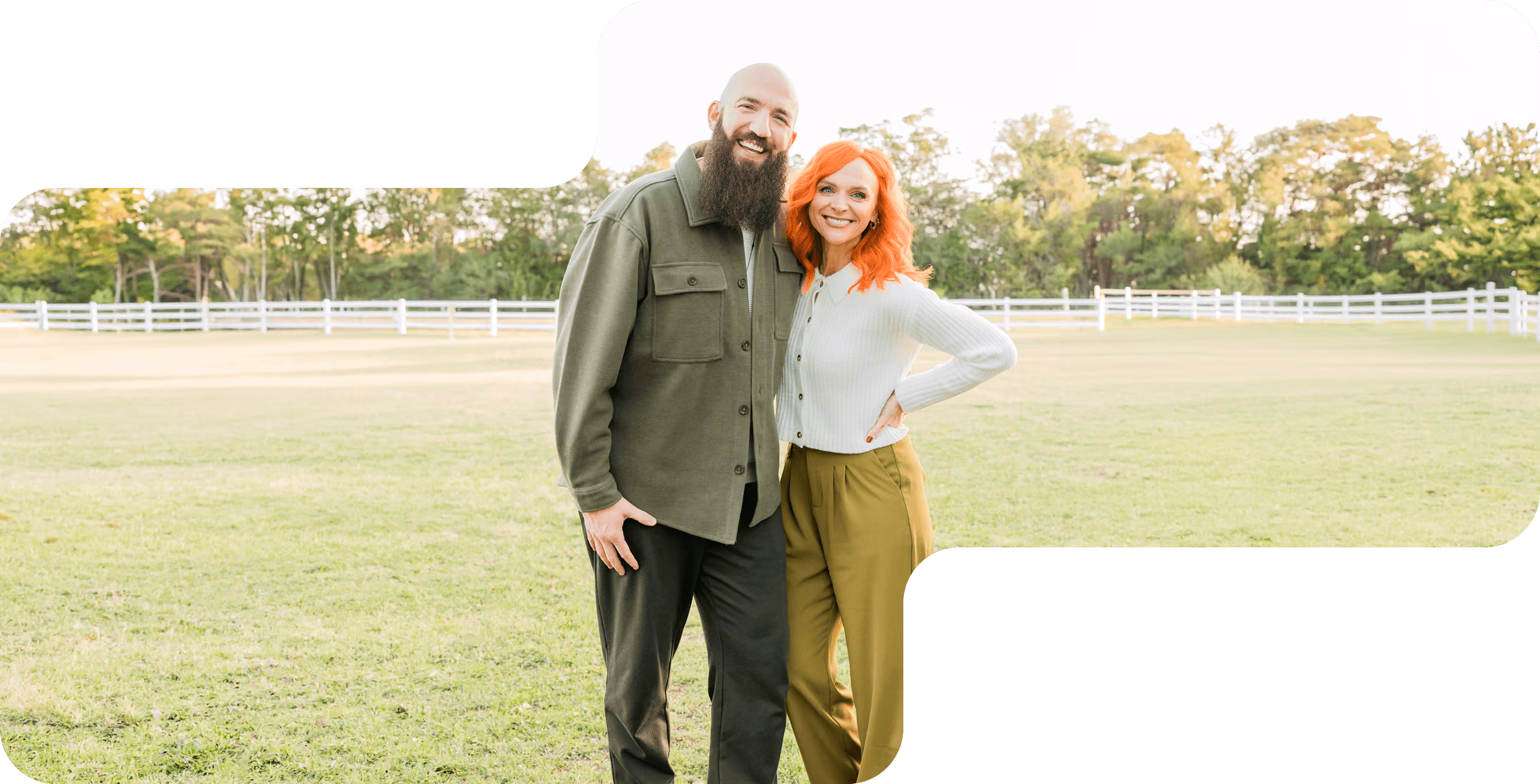Smiling bald man with beard and woman with bright orange hair standing outdoors in a green park.