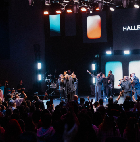 Live worship band performing on stage with audience watching under colorful lights and screen text.
