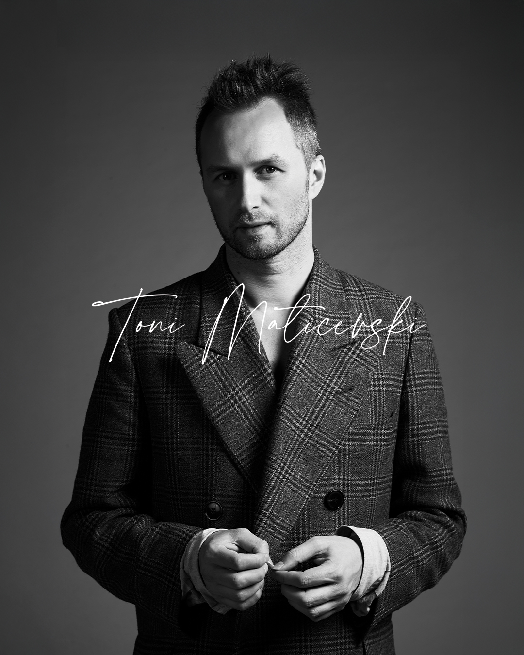 Toni Maticevski in a checkered coat buttoning it, against a plain dark background.