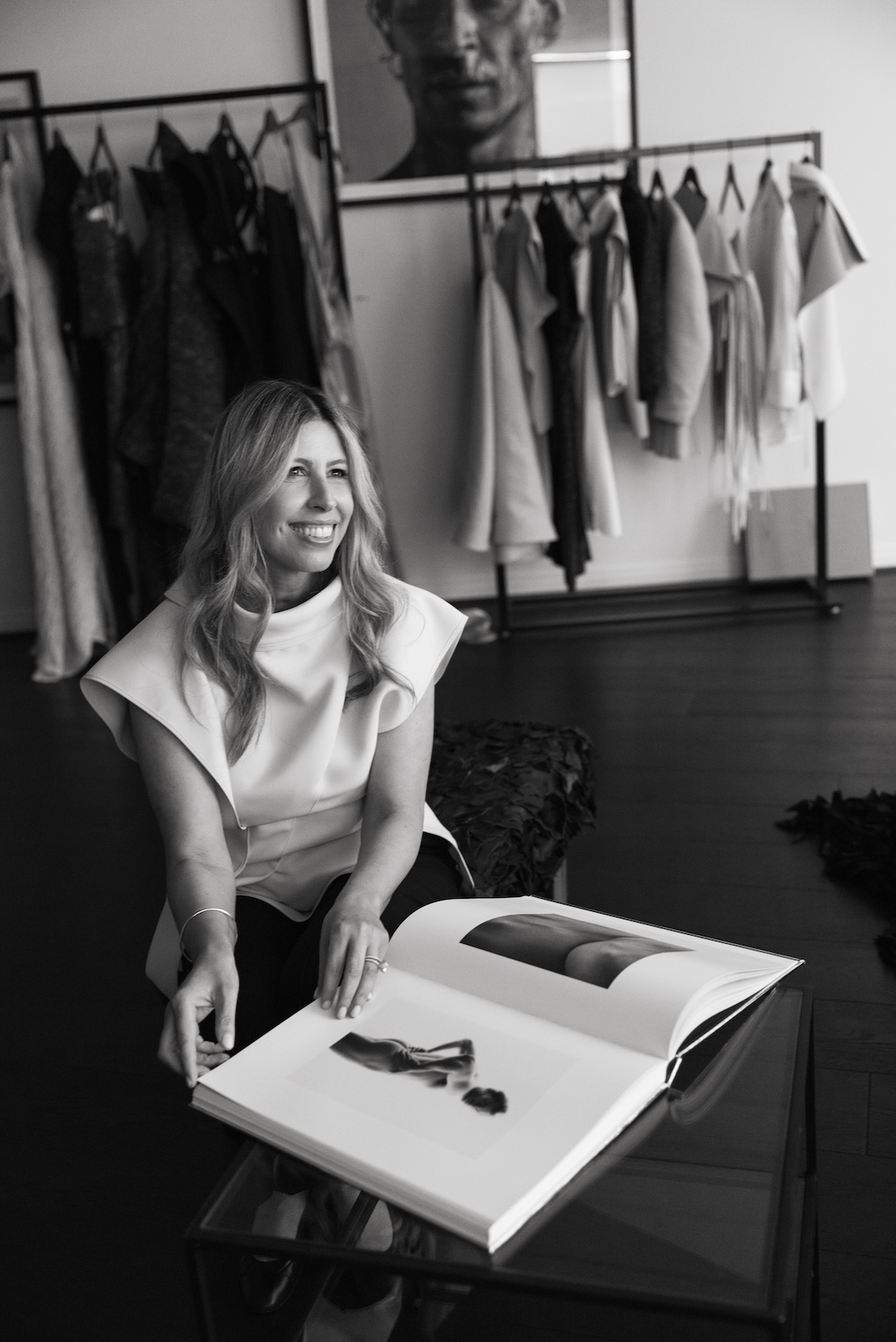 Helen Mantzis sitting, looking away, with a large photo book open on a glass table in front of her.