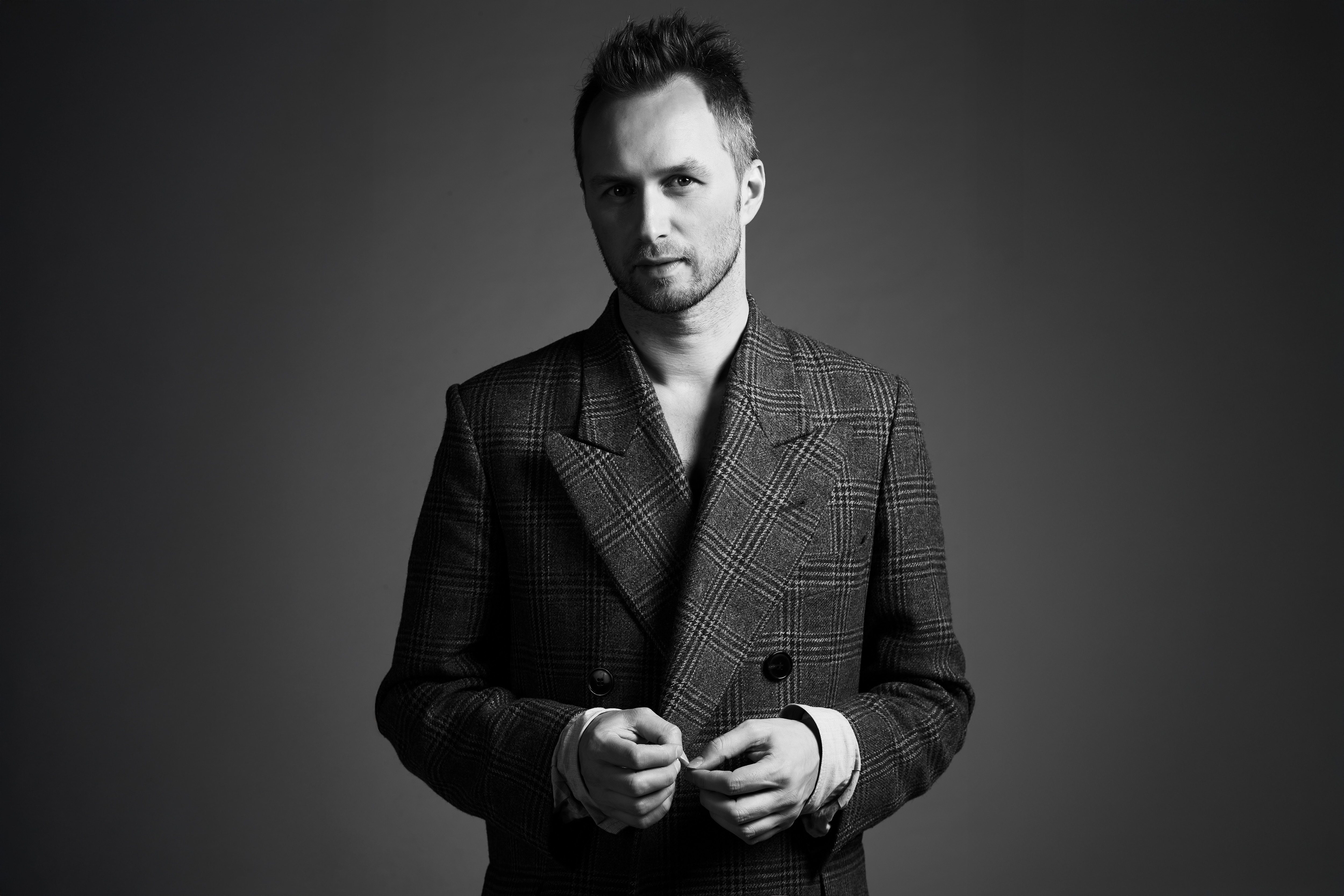 Toni Maticevski in a plaid double-breasted blazer looking at the camera with hands adjusting the jacket.
