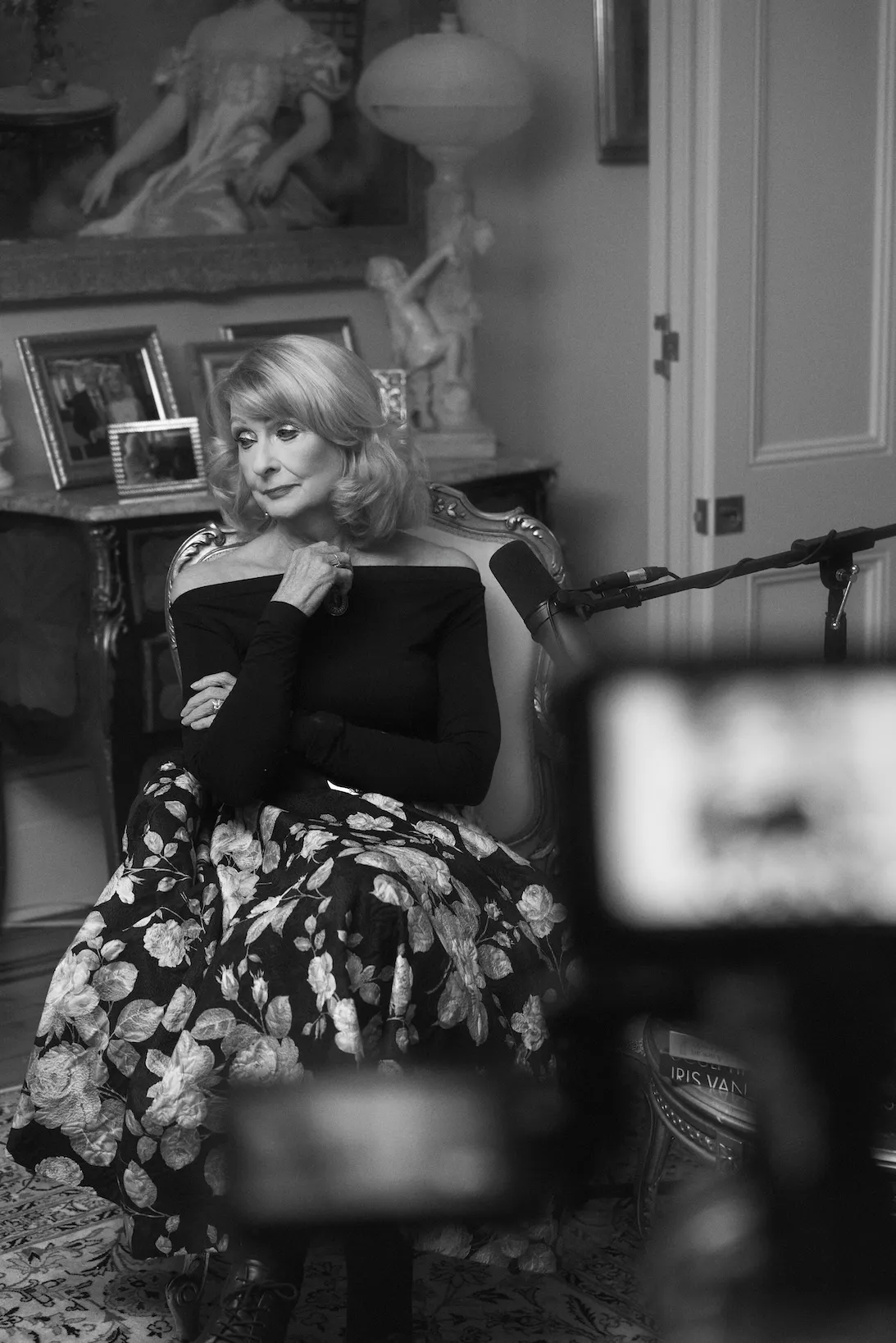 A Krystyna Campbell Pretty AM in a floral skirt and black top poses thoughtfully in a vintage room with framed photos.