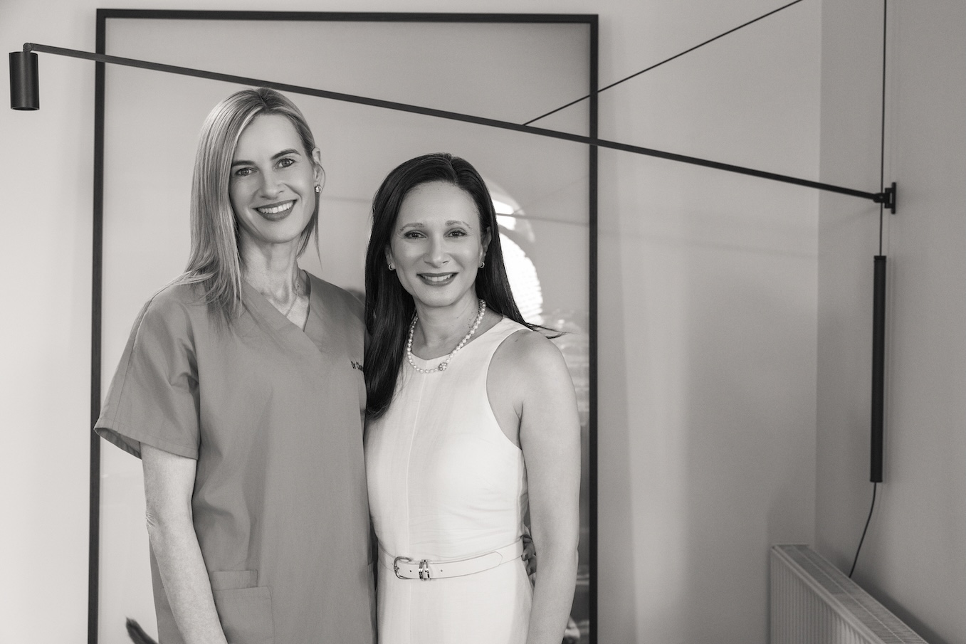 Krystyna Campbell-Pretty AM & Helen Mantzis in elegant black outfits stand in a vintage room with framed paintings behind them.