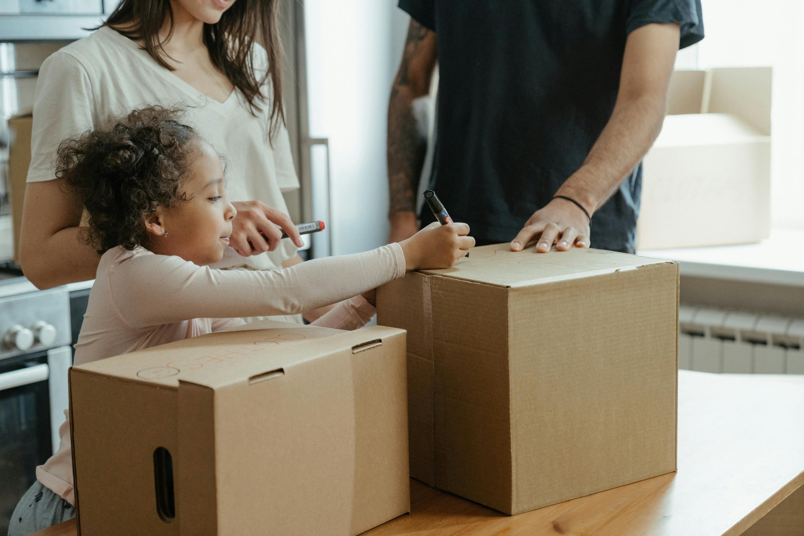 A young child writing on a cardboard box with an adult and another person assisting nearby during packing.