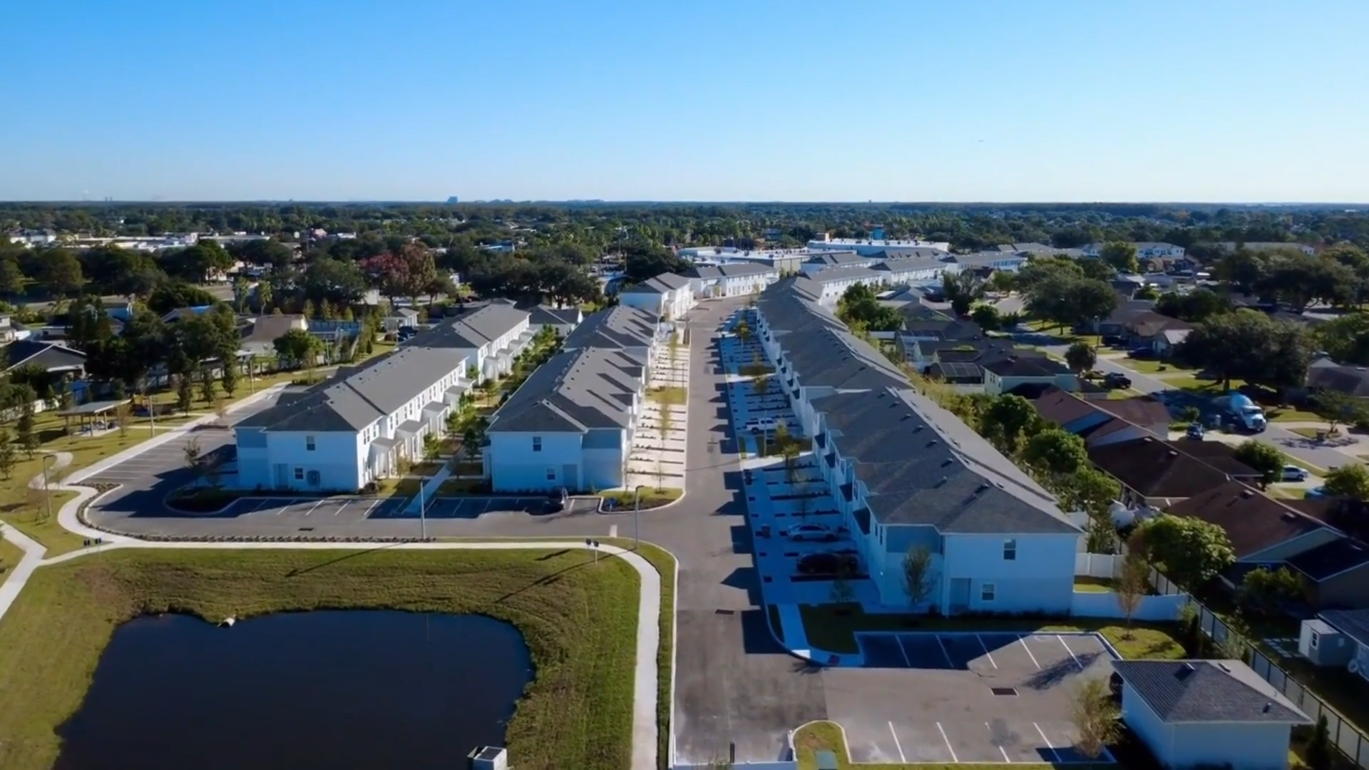 Aerial view of a residential neighborhood with rows of townhouses, parking lots, and a small pond under a clear blue sky.