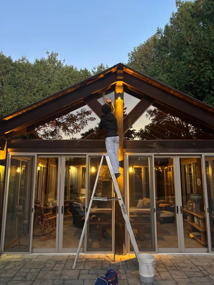 Person on ladder cleaning or reaching towards an outdoor wall light on a wooden patio structure at dusk.