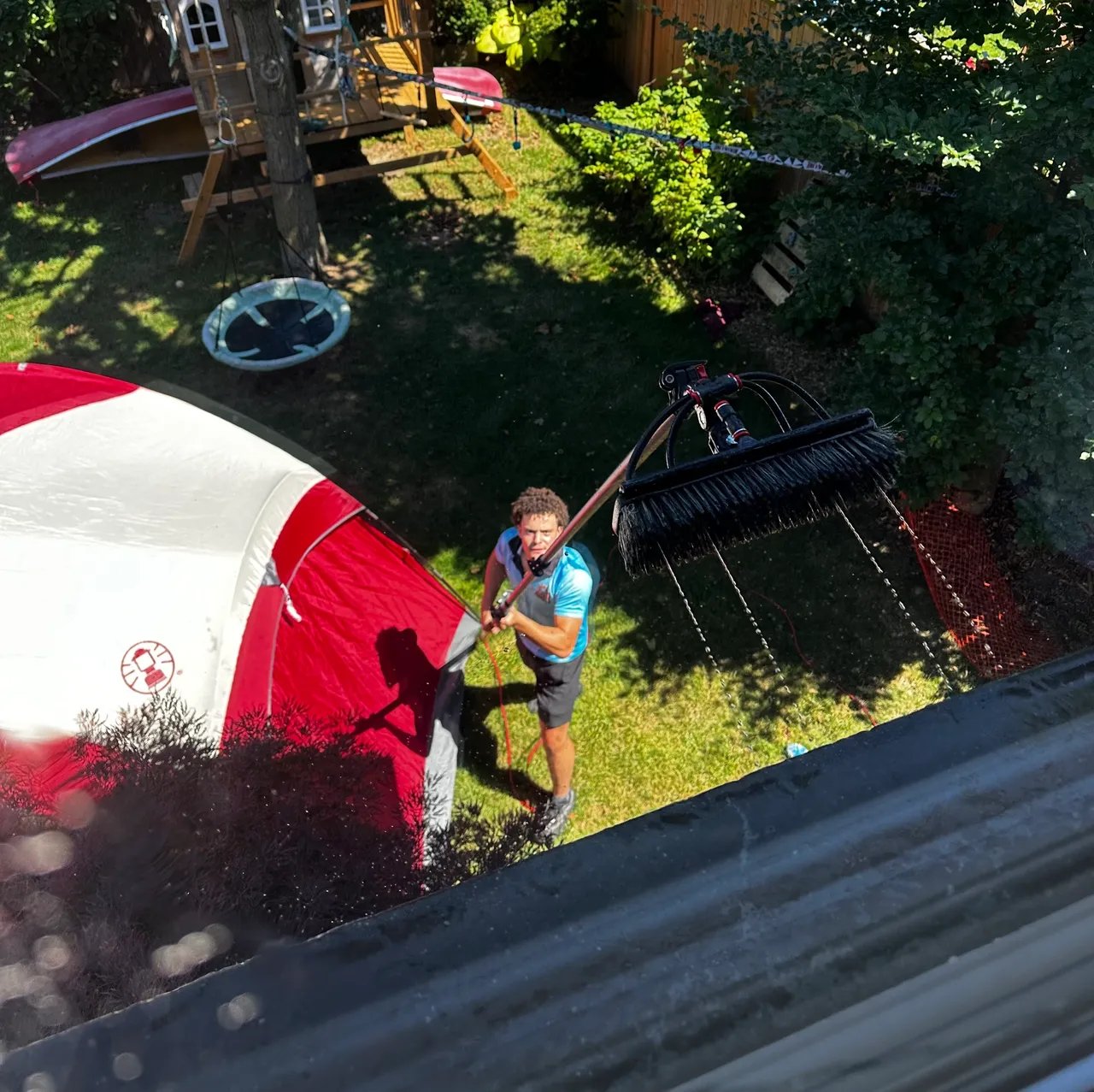 Man cleaning second-story windows using an extended water-fed brush in a backyard.