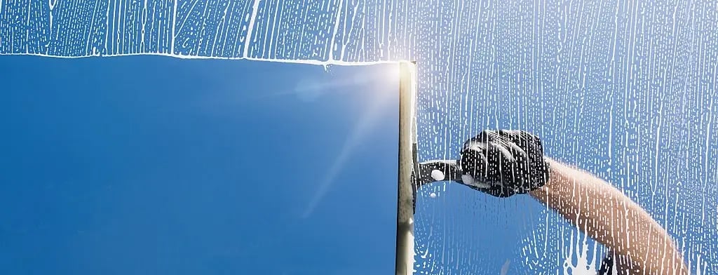 A person wearing a black glove cleaning a window with a squeegee against a clear blue sky.