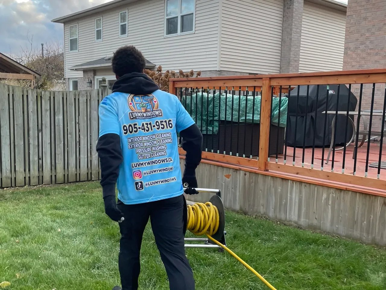 Person wearing a blue LuvMyWindows shirt walking on grass, holding a yellow water hose reel in a residential backyard.