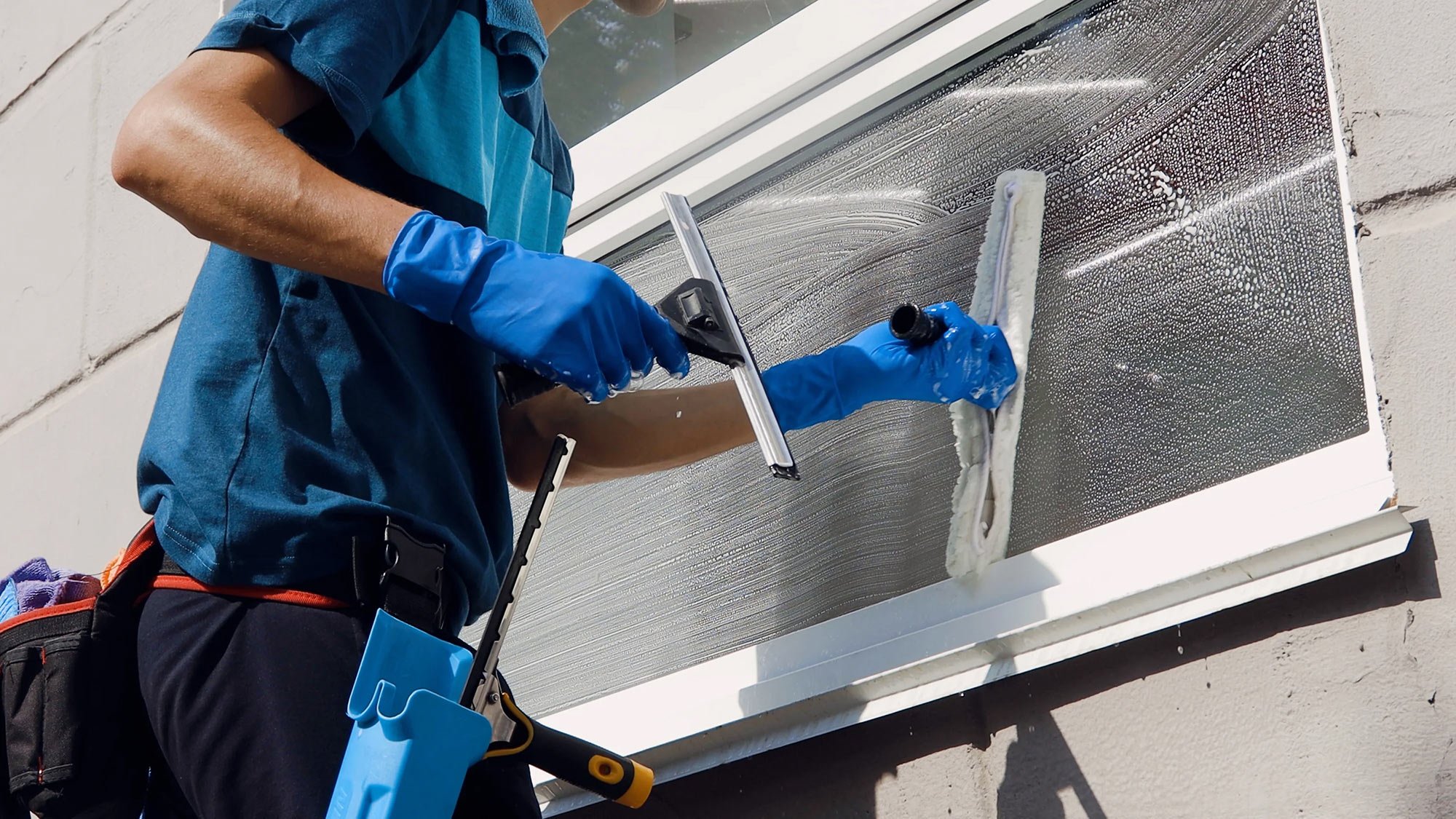 Person wearing blue gloves cleaning a large window with a squeegee and a window scrubber.