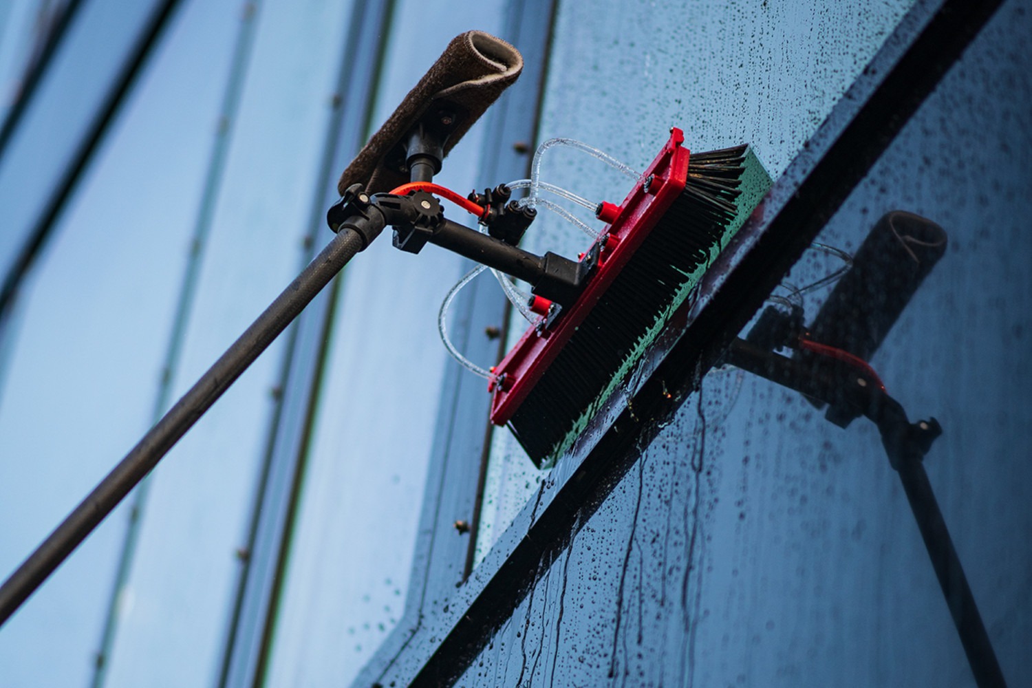 Long-handled brush with water tubes cleaning a wet glass window on a building exterior.