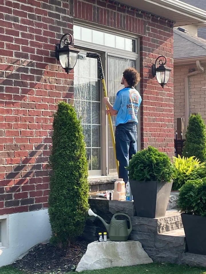 Man wearing a blue shirt cleaning a large front window of a brick house using an extended window cleaning tool during daytime.