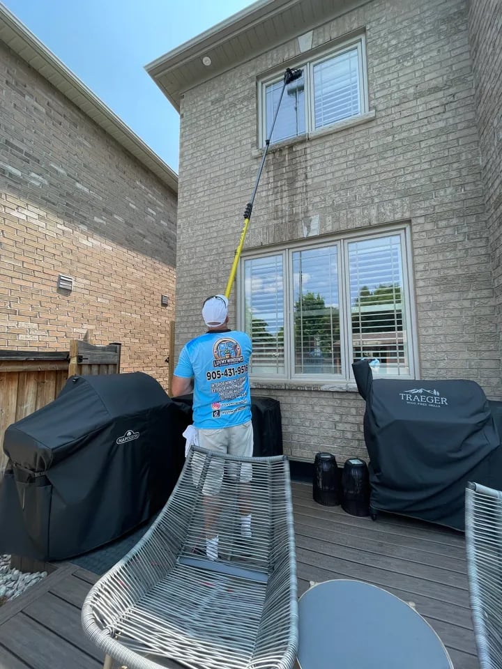 Person wearing a blue shirt and white cap cleaning an upstairs window of a brick house using a long pole with a cleaning attachment.