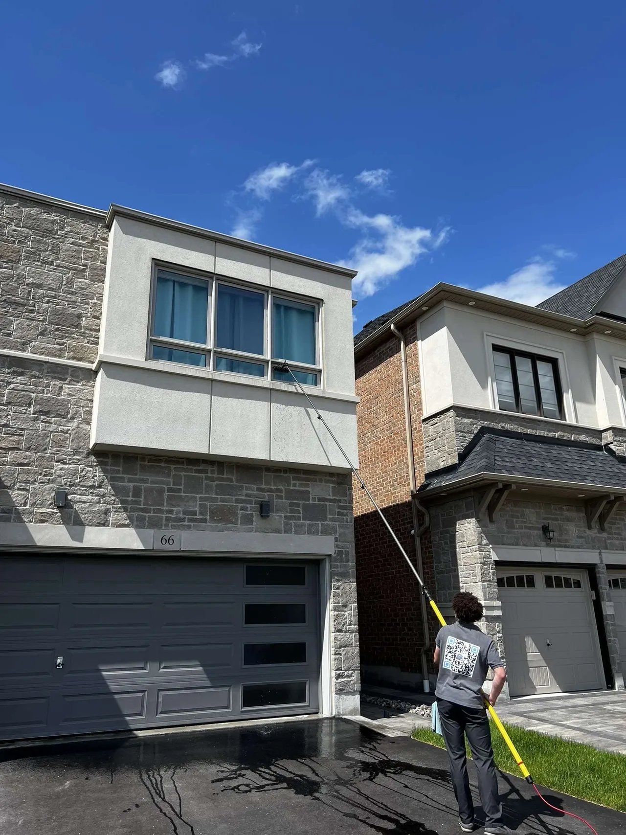 Person cleaning second-story window of a modern house using a long pole with a brush attachment under a blue sky.