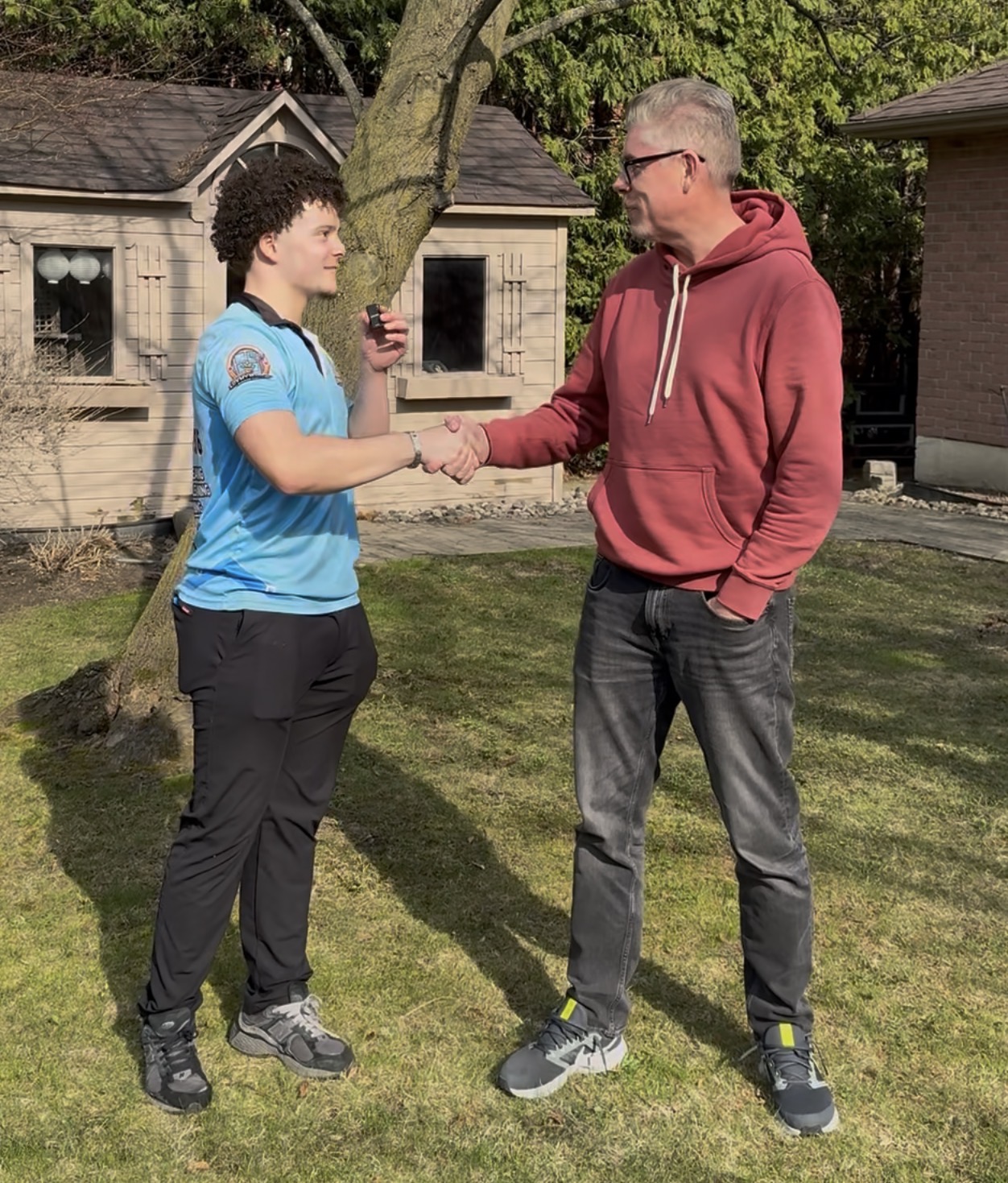 Two people shaking hands outdoors with a house blurred in the background.