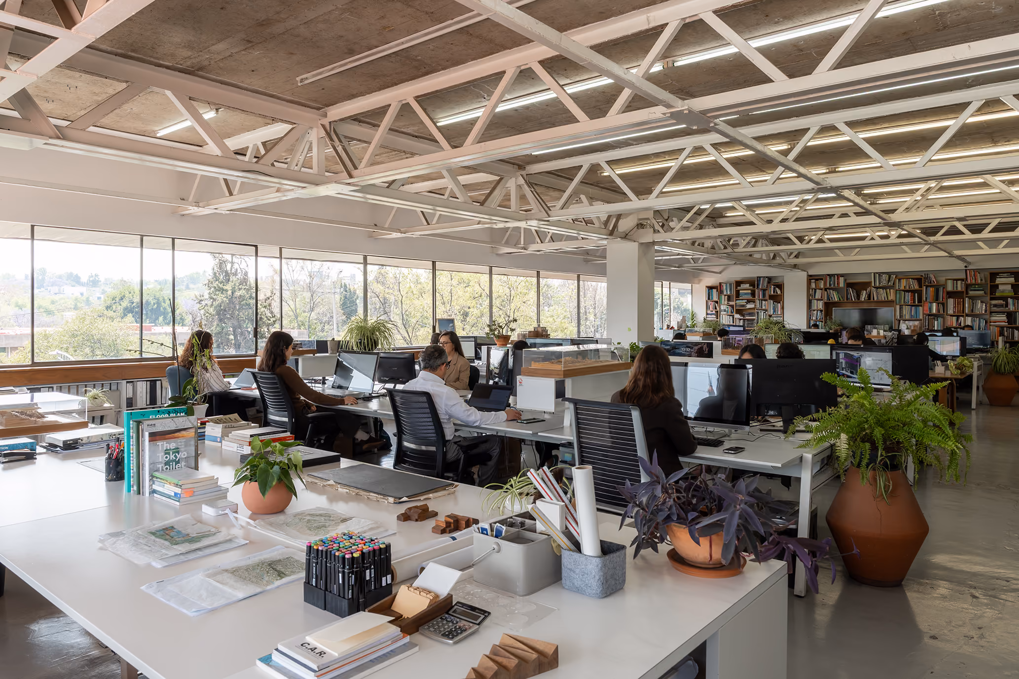 Estudio de a|911: espacio de trabajo abierto con mesas, estructura de acero en el techo y ventanales hacia los árboles