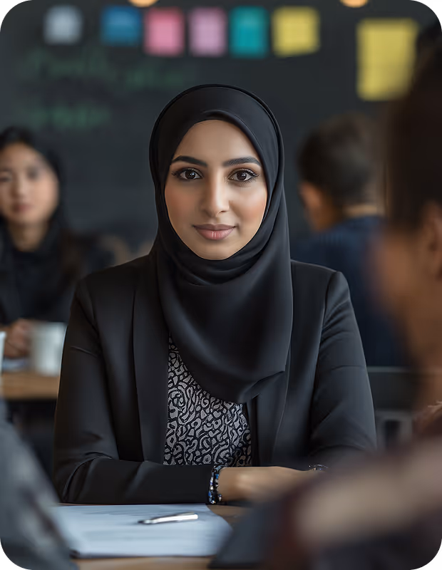 Confident woman wearing a black hijab and blazer sitting at a table with documents and a pen.